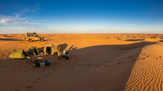 Vue panoramique d'un campement dans les dunes du Sahara au coucher du soleil avec équipement d'expédition et 4x4